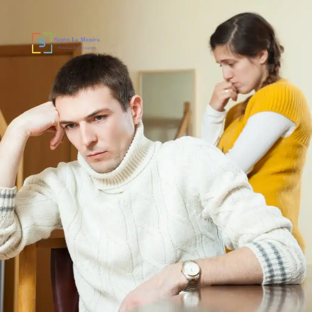 Coppia seduta in casa in un momento di tensione emotiva, con l’uomo pensieroso in primo piano e la donna preoccupata sullo sfondo.