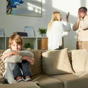 Bambino seduto sul divano con aria triste mentre due adulti discutono animatamente sullo sfondo, simbolo di conflitti familiari.