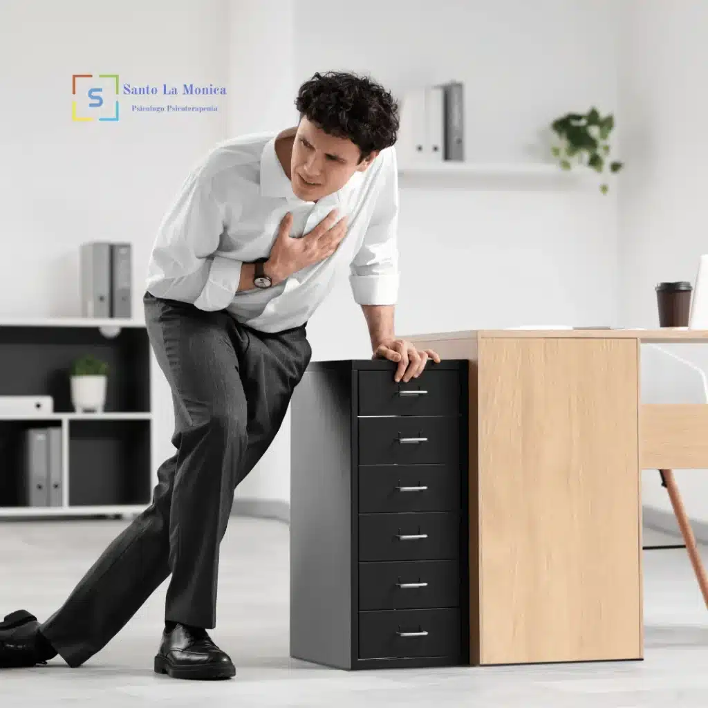 Uomo in ufficio si appoggia a un mobile con una mano al petto, visibilmente in difficoltà e con respiro affannoso.